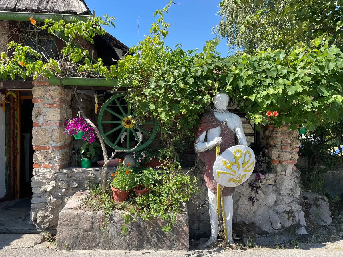 Ulaz u Restoran Sojenica — kameni zid, loza i skulptura ratnika sa štitom